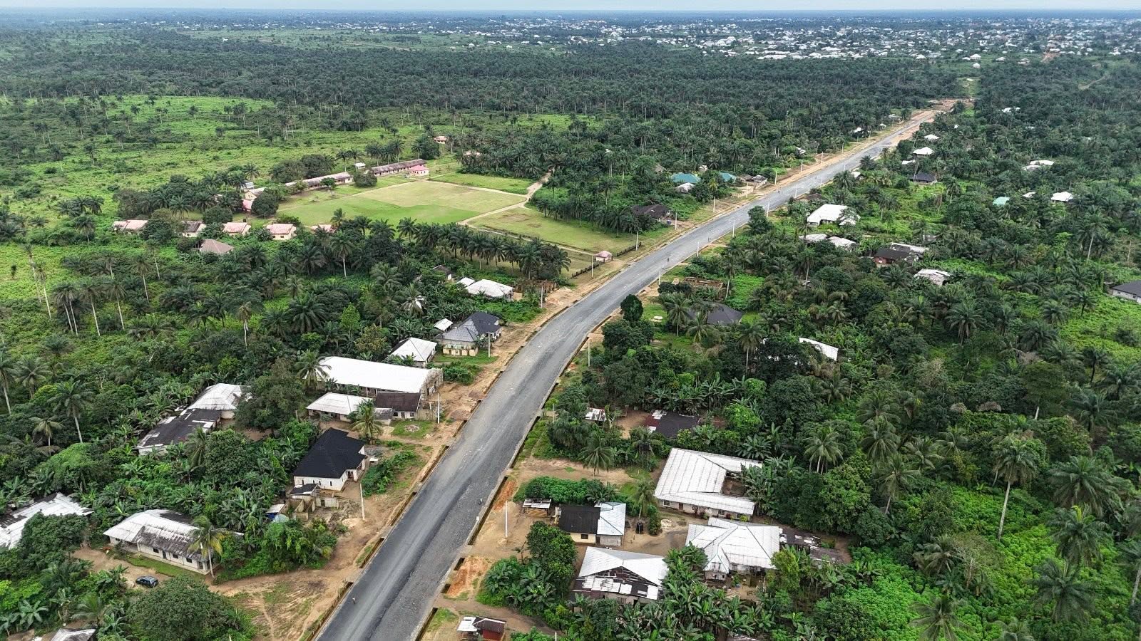Wide drone view of the straight asphalt carriageway running past farmland and a school field